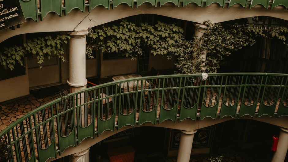 A view of a multi-level interior courtyard featuring a curved green metal railing along the edge of an upstairs walkway. Beneath the balcony, there are lush green plants with broad leaves growing alon